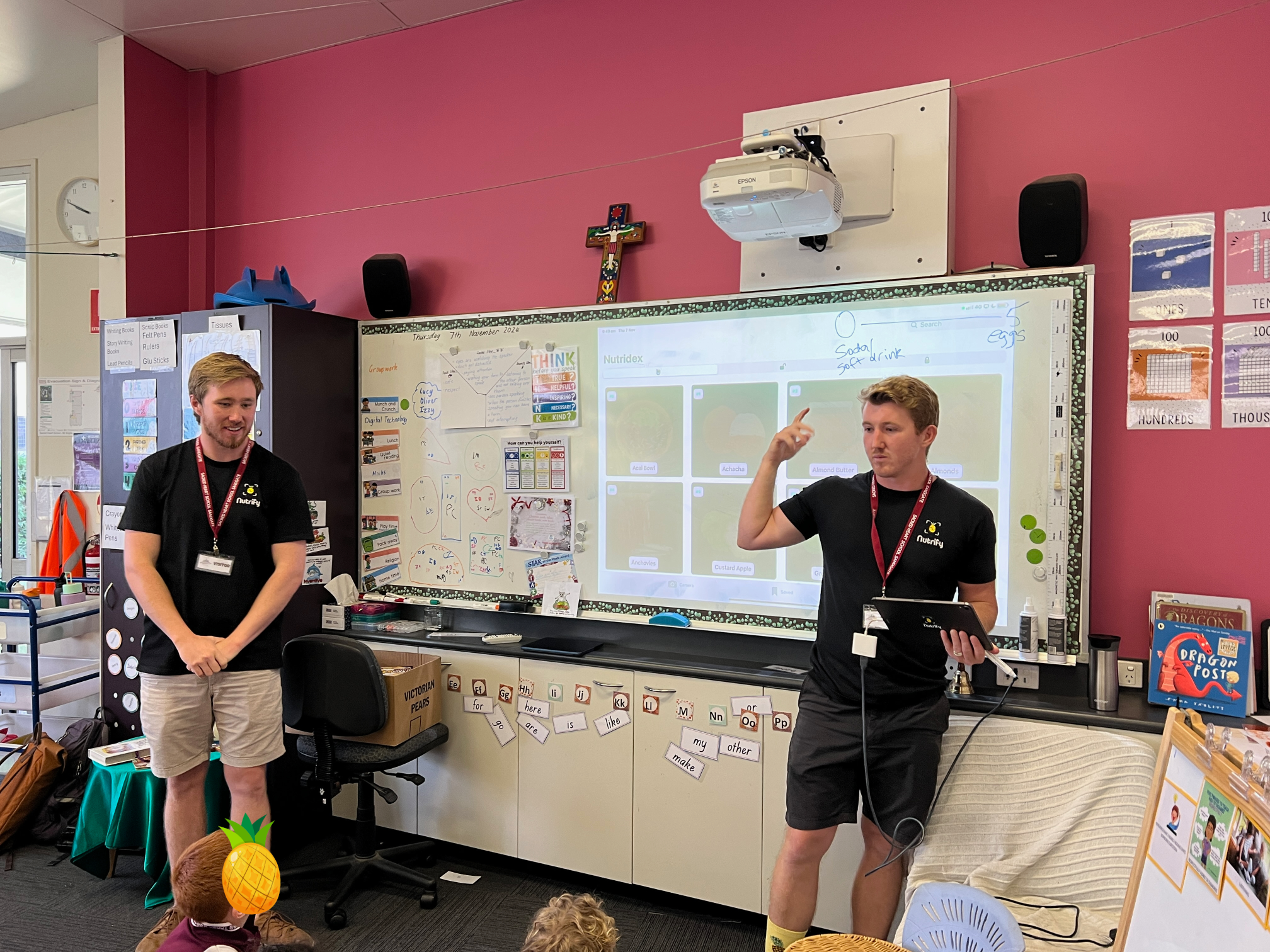 Two presenters from Nutrify stand in a classroom in front of a whiteboard and projector screen, engaging with students seated on the floor.