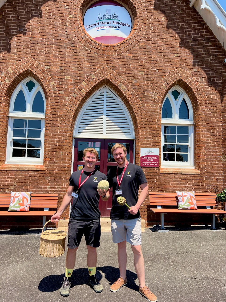 Two men wearing black Nutrify shirts and lanyards stand in front of Sacred Heart Sandgate, a brick building with arched windows. One holds a basket, and the other holds a melon and an artichoke. They are smiling, and there are benches with colorful cushions on either side of them.